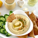 Homemade Hummus in white bowl with pita chips and cucumber slices on side