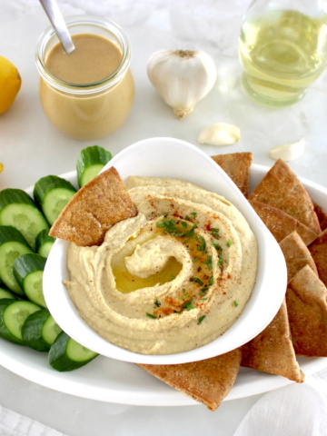 Homemade Hummus in white bowl with pita chips and cucumber slices on side