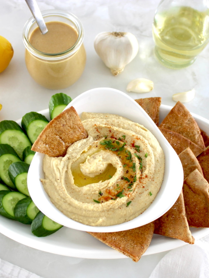 Homemade Hummus in white bowl with pita chips and cucumber slices on side