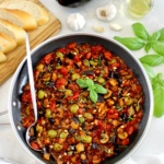 Eggplant Caponata in skillet with silver serving spoon on side and fresh basil garnish