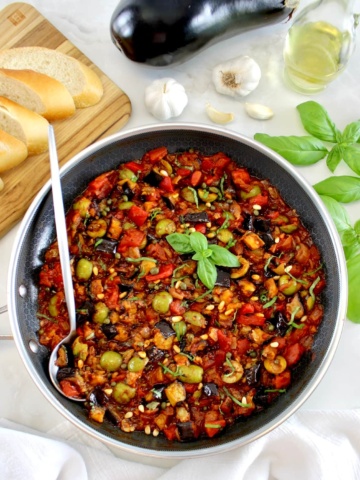 Eggplant Caponata in skillet with silver serving spoon on side and fresh basil garnish
