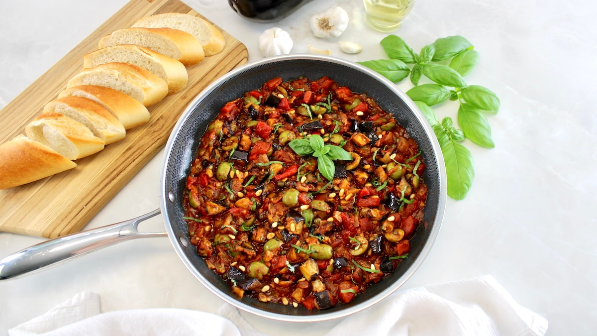 Eggplant Caponata in skillet with sliced baguette and basil in back