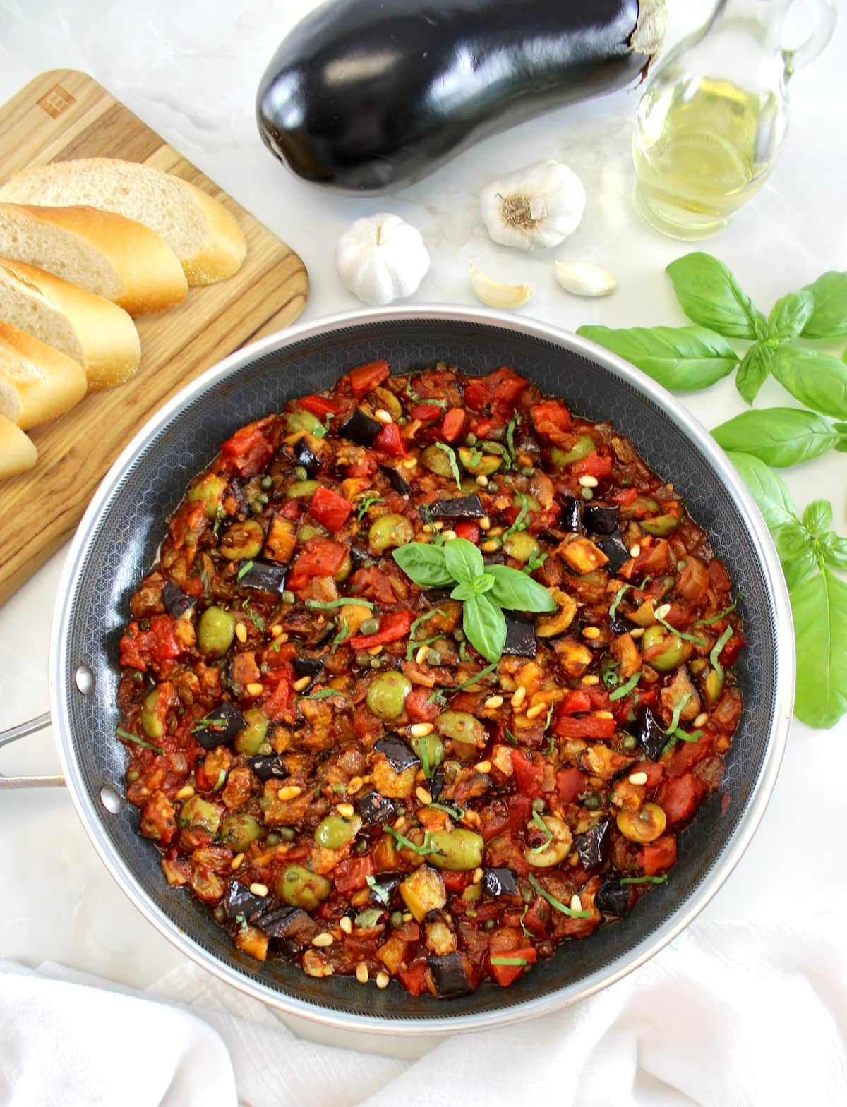 overhead view of Eggplant Caponata in skillet with sliced baguette, eggplant and basil in back