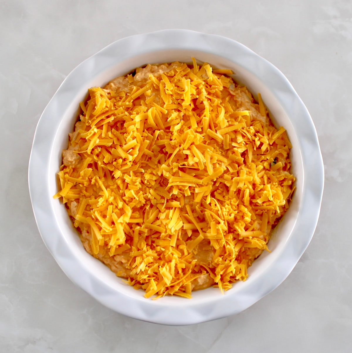 Cottage Cheese Buffalo Chicken Dip in white pie dish with shredded cheddar on top unbaked
