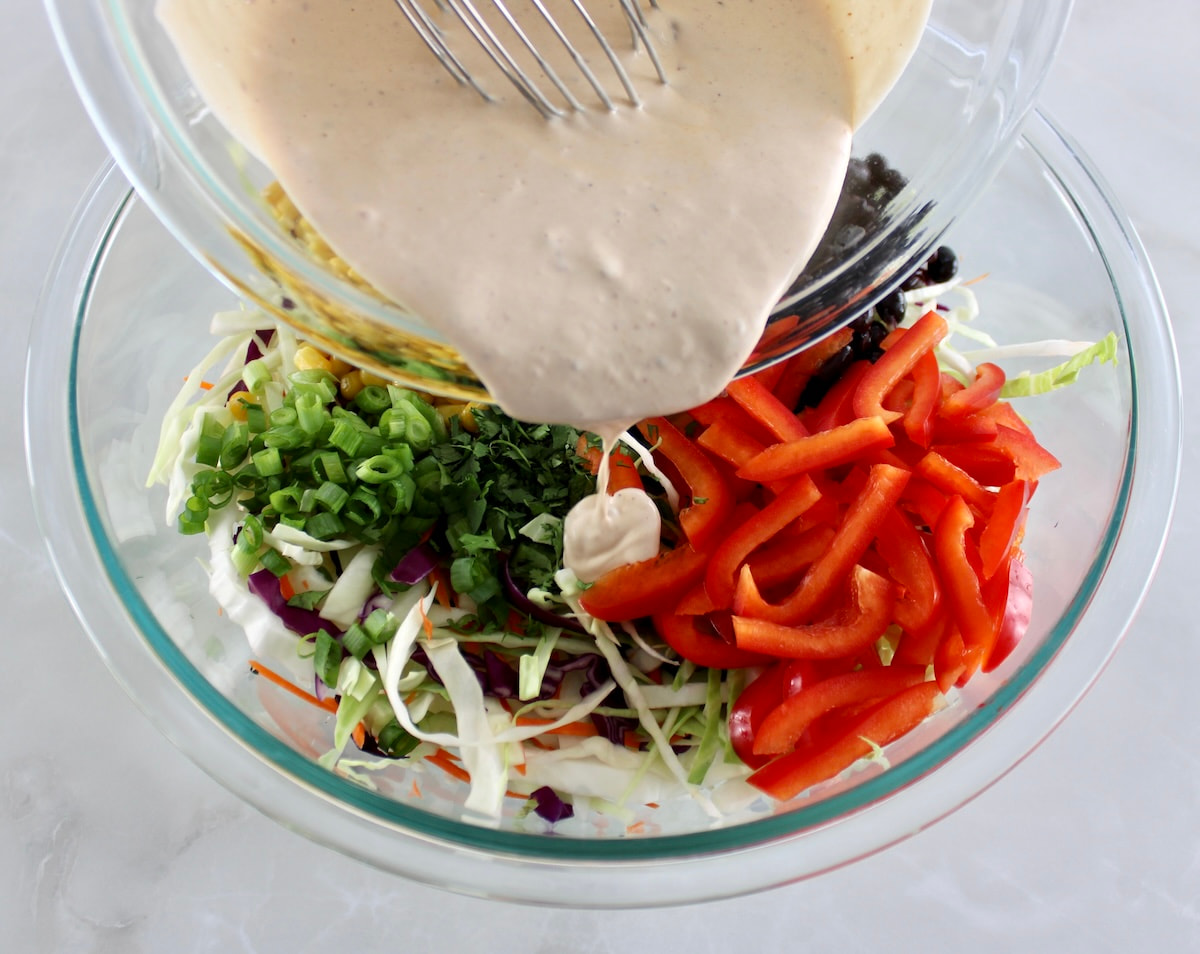 Cowboy Coleslaw ingredients in glass bowl unmixed with dressing being poured over the top
