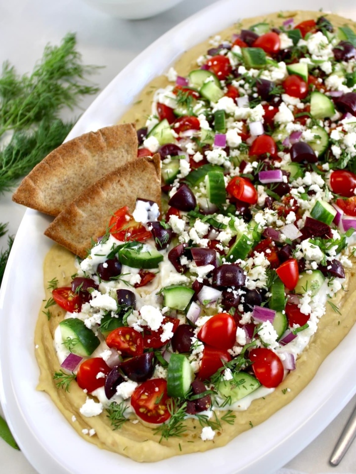 Layered Greek Hummus Dip on oval white platter with 4 pita chips on side