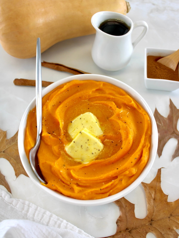 Mashed Butternut Squash in white bowl with 2 pats of butter and spoon on side